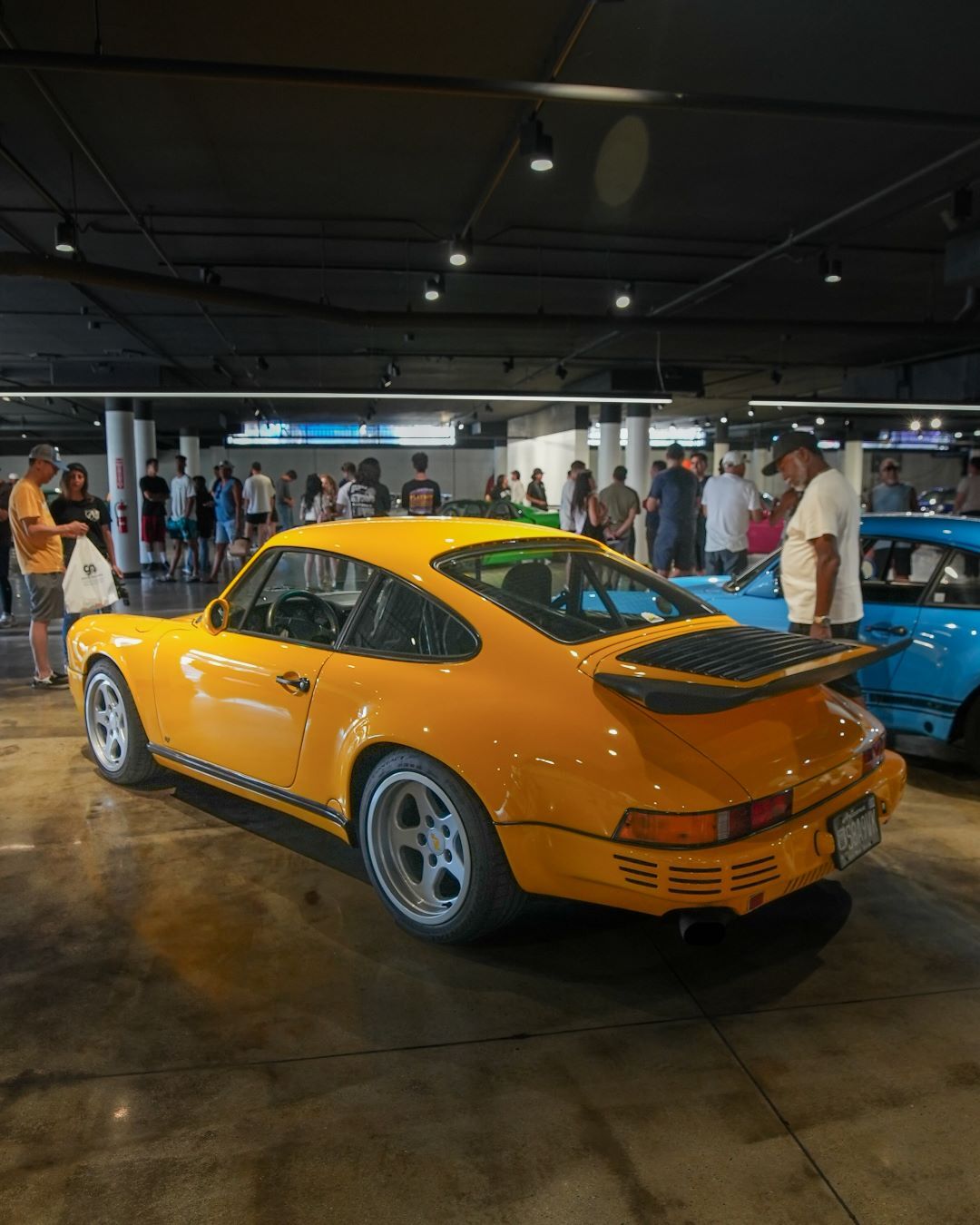A bright yellow vintage Porsche 911 with a rear spoiler sits indoors, surrounded by a crowd of people.