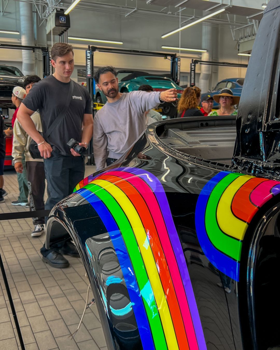 People admiring a black car with vibrant rainbow stripes in a garage, auto lifts and other cars in the background.