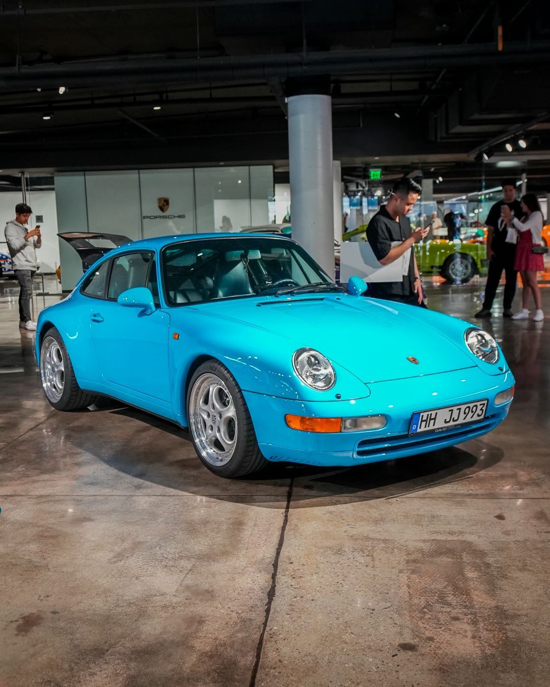 A bright blue Porsche with shiny wheels parked on a concrete floor in a dealership, with people nearby.