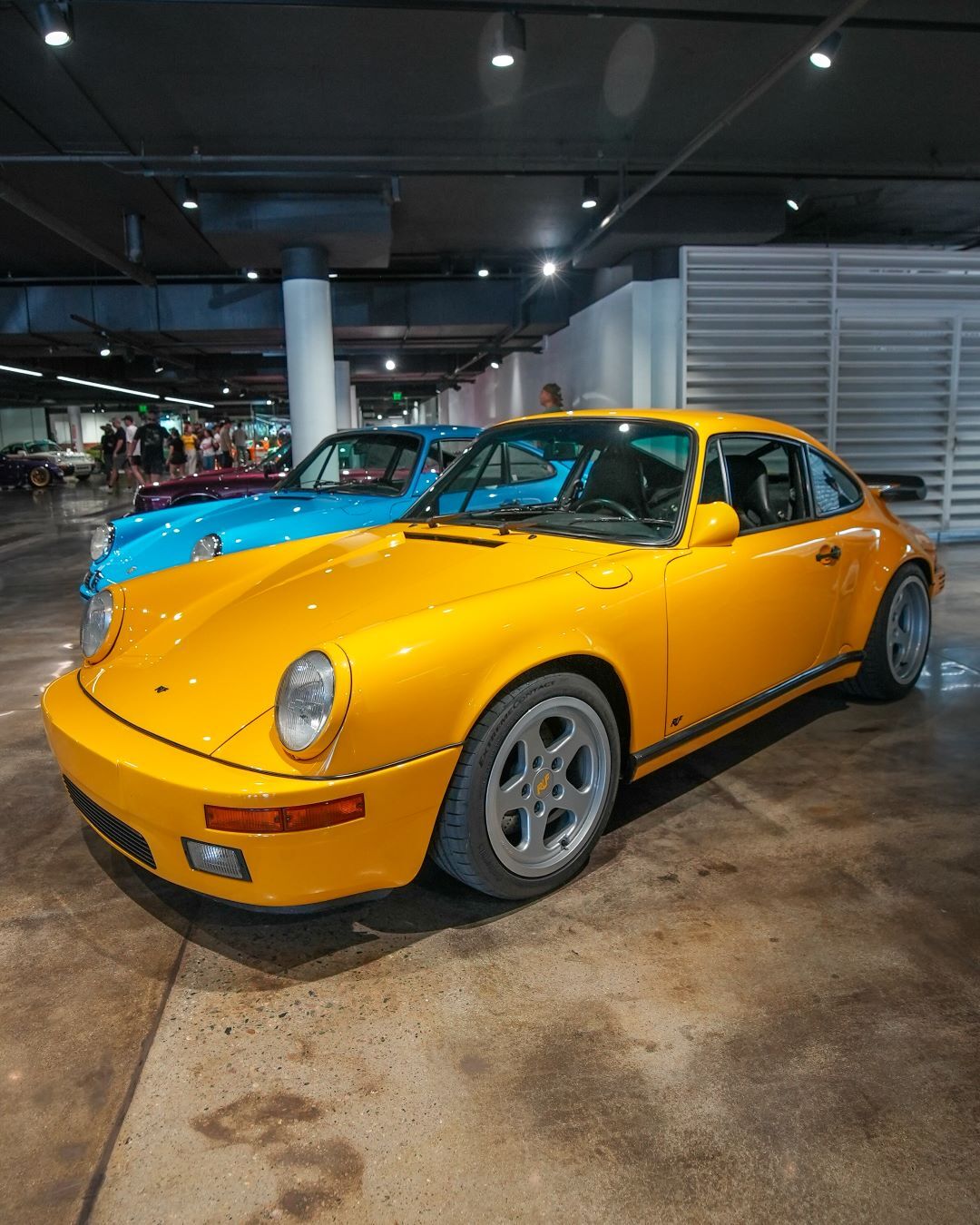 A bright yellow Porsche 911 in the foreground, with a blue classic Porsche 911 behind it in an indoor parking lot.