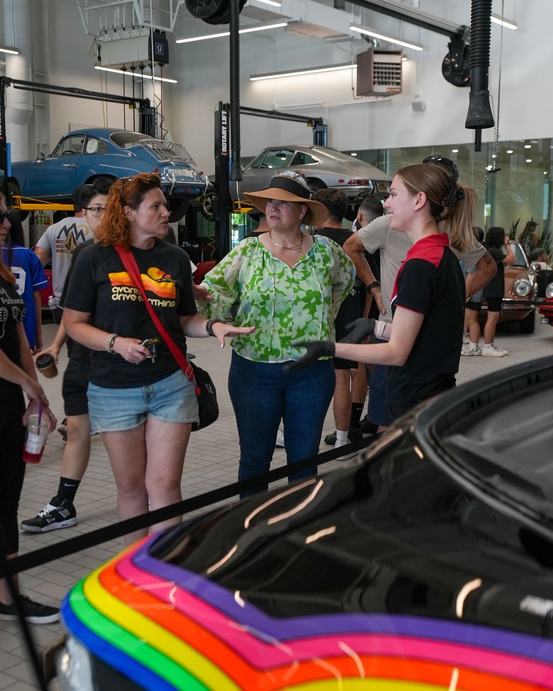 People chatting in a garage with classic cars on lifts and a black car with a rainbow stripe.