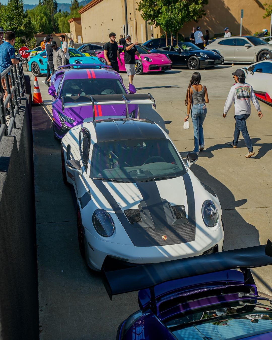 A white Porsche with black racing stripes and a purple Porsche are the closest in a parking lot filled with colorful sports cars.