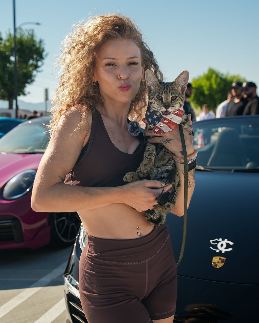 A woman with curly hair pouts while holding a serval cat in an American flag bib, standing by a black Porsche.