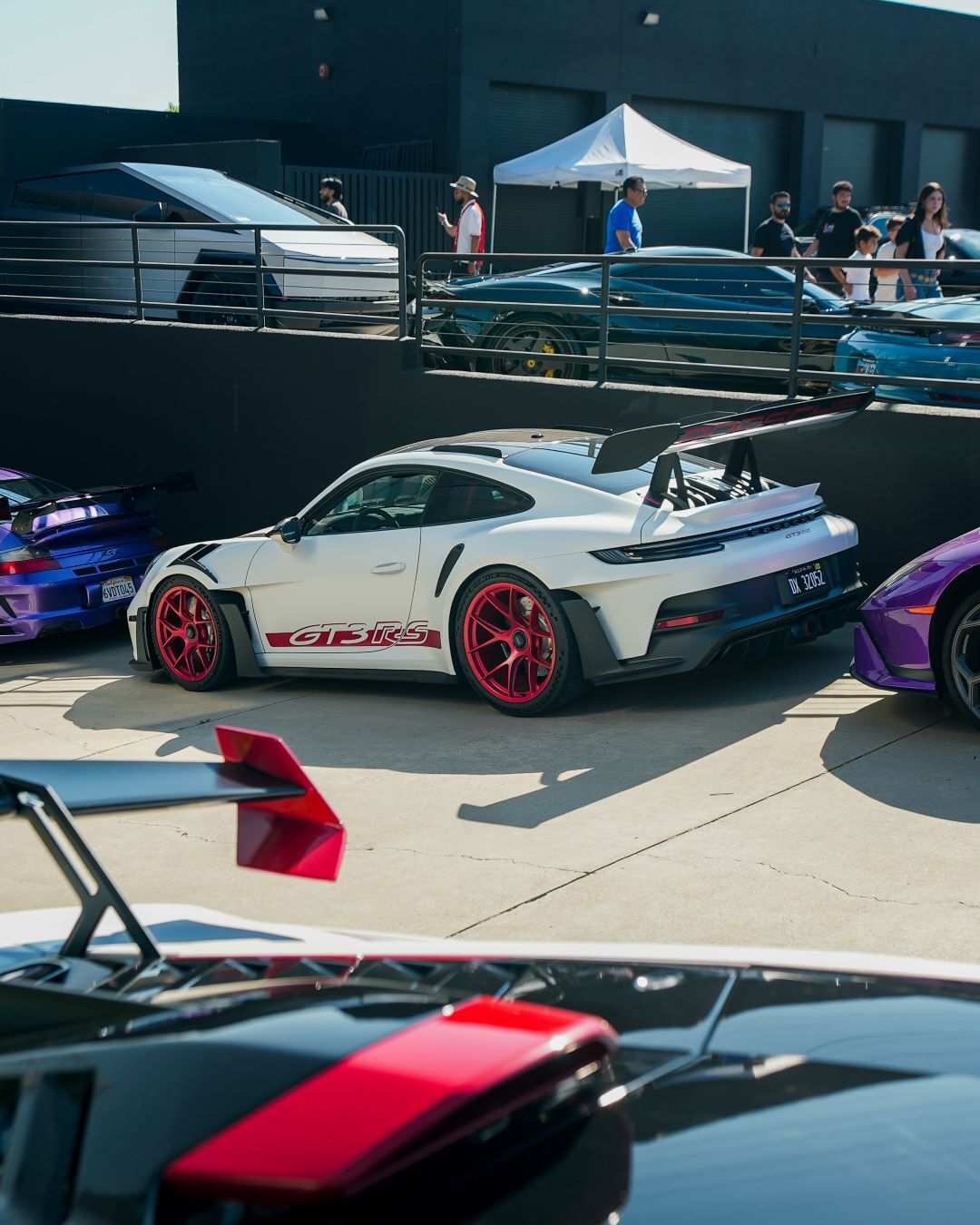 White Porsche GT3RS with red rims and large spoiler, parked among other luxury cars at an outdoor event.