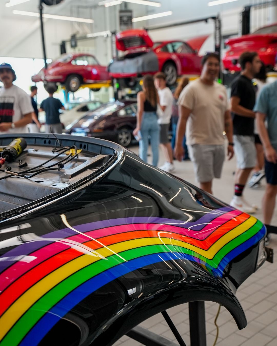 Front panel of a black classic car with bright rainbow-colored stripes, people and red cars in a garage background.