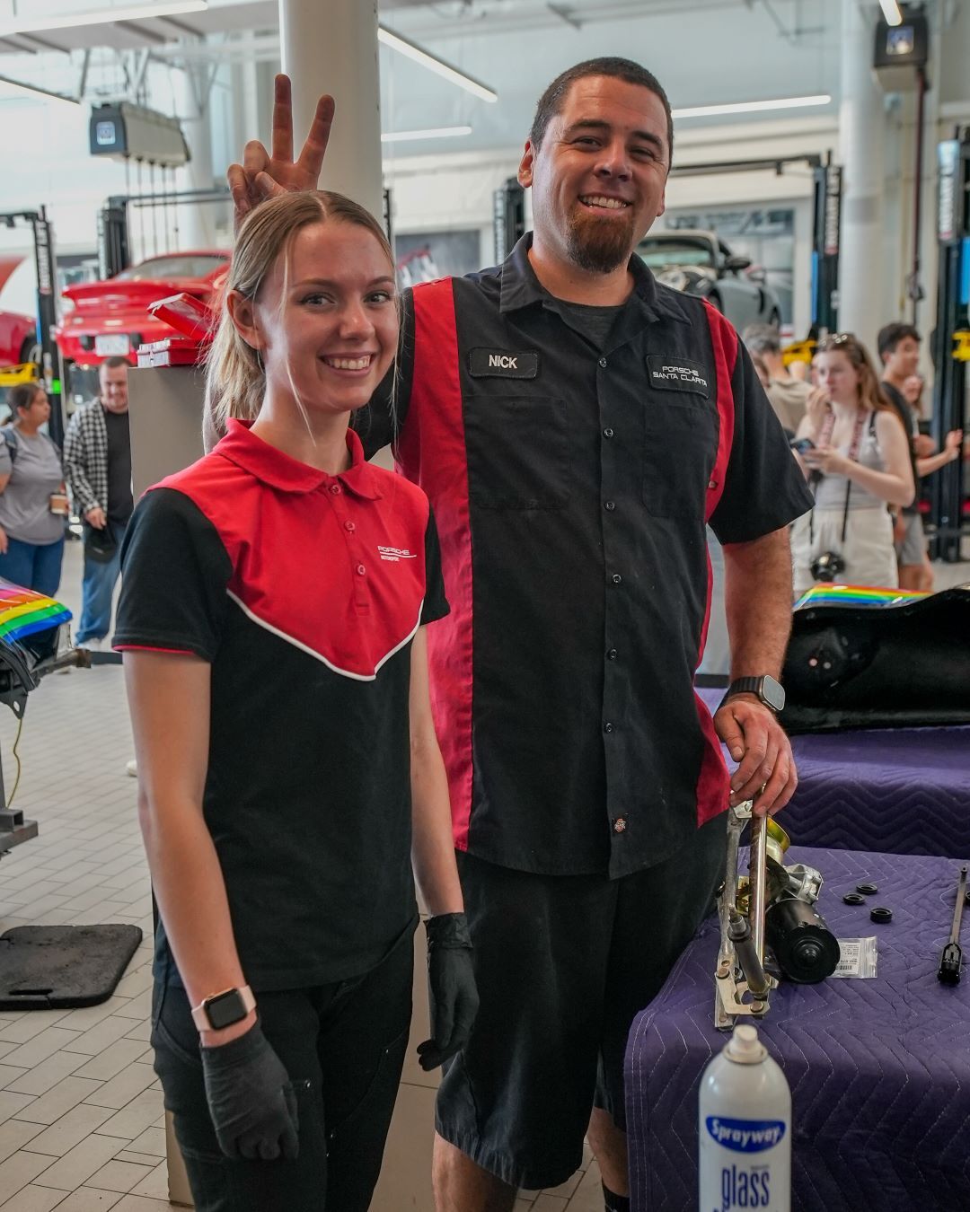 Two smiling auto technicians in a garage, one giving peace sign behind the other's head.