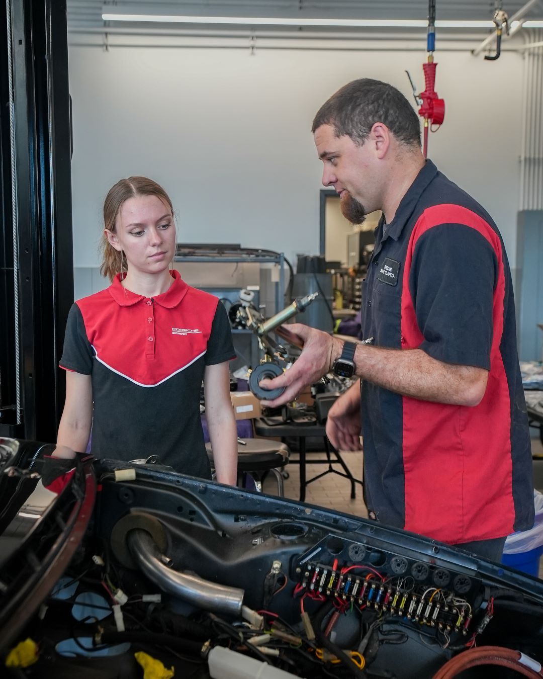 Mechanic mentor explaining a part to a female student over an open car engine in a garage.