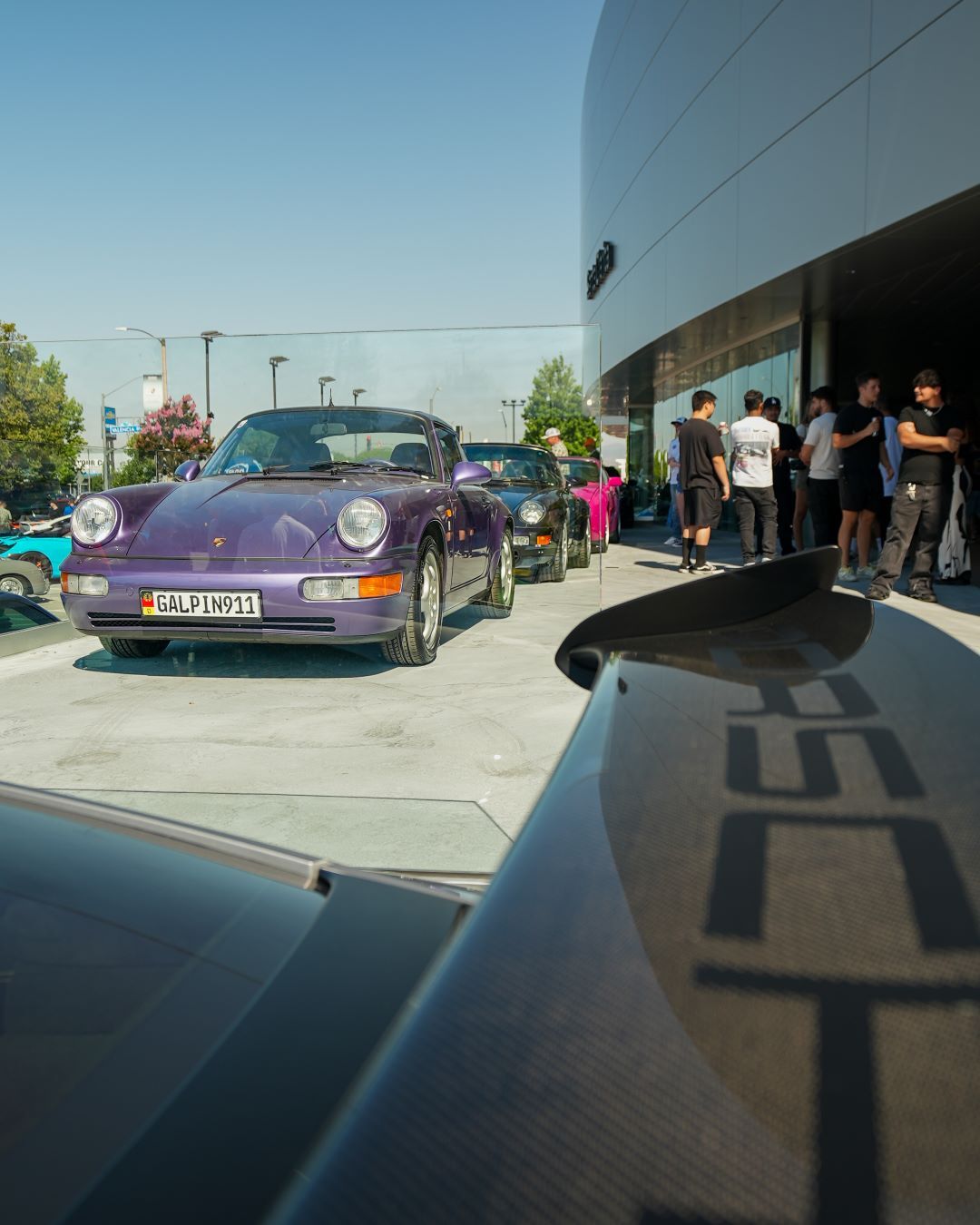 A lineup of classic Porsche sports cars, including a prominent purple one, attracting a crowd outside a modern building.
