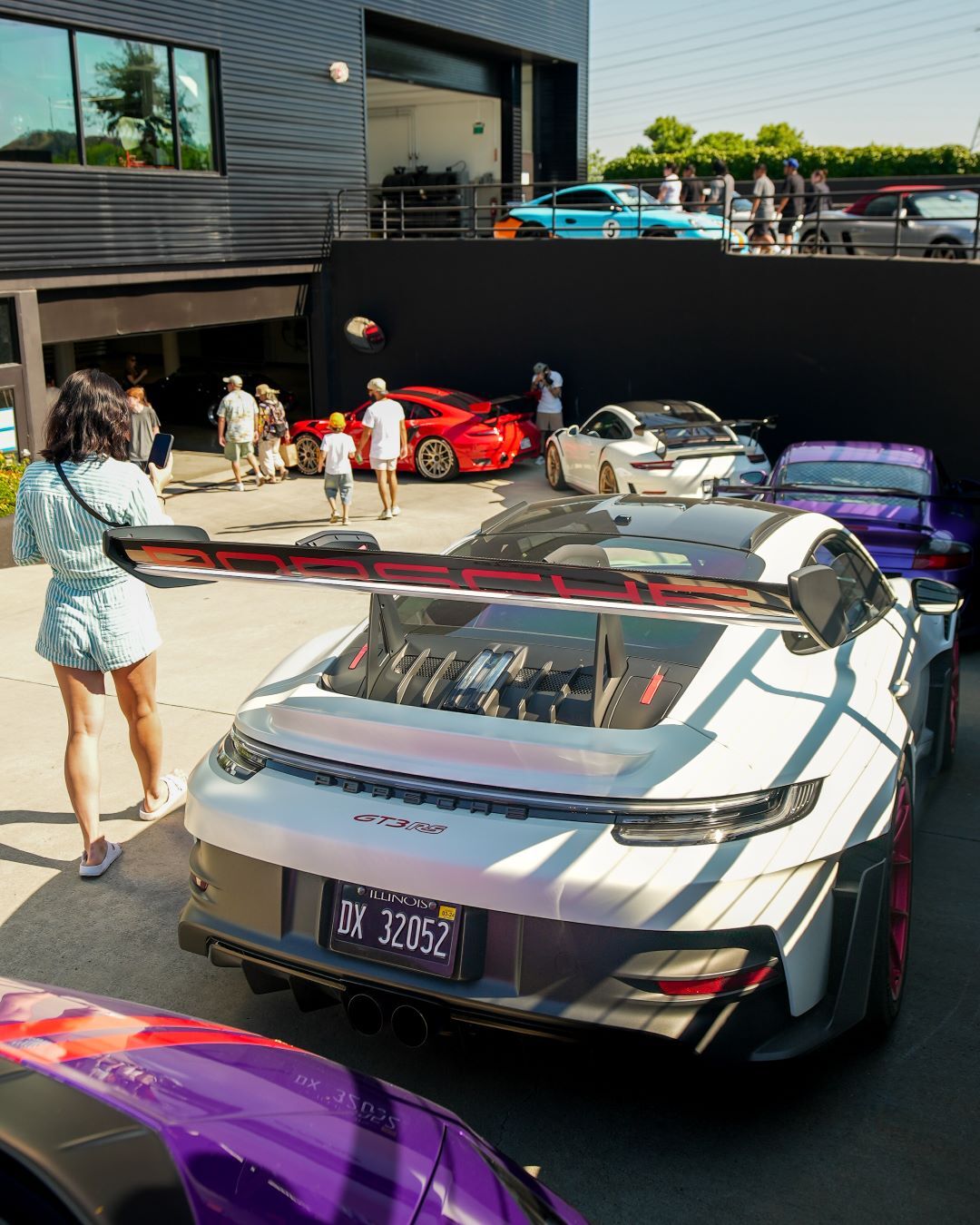 Rear view of a white Porsche GT3 RS with a large spoiler, surrounded by other luxury cars and people.