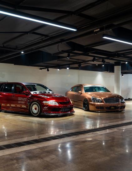 Two customized cars, one red and one bronze, are parked inside a well-lit garage.