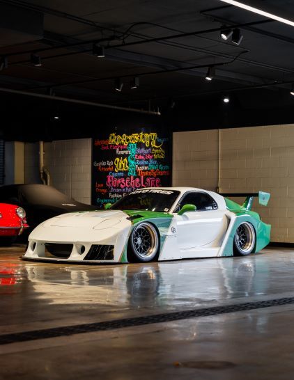 A white and teal race car with a large spoiler sits in a dimly lit garage with a mural on the wall.
