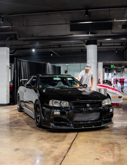 A man in a gray shirt and baseball cap polishes a black Nissan Skyline in a modern garage.