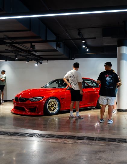 Three people examine a red BMW with gold wheels in a brightly lit parking structure.