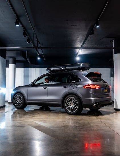 A dark gray Porsche Cayenne SUV with roof rack sits in a dimly lit parking garage.