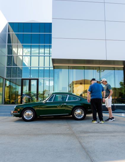 Three people look at a vintage green Porsche parked in front of a modern glass building.