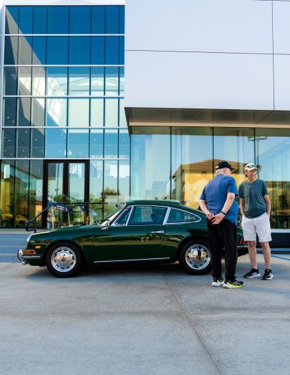 Three men converse beside a classic green Porsche parked in front of a modern glass building.