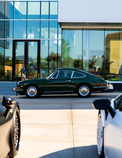 A classic green Porsche sits parked in front of a modern glass building.
