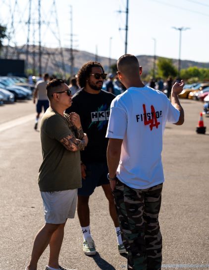 Two men in shorts and sunglasses converse in a parking lot with a crowd and cars behind them.