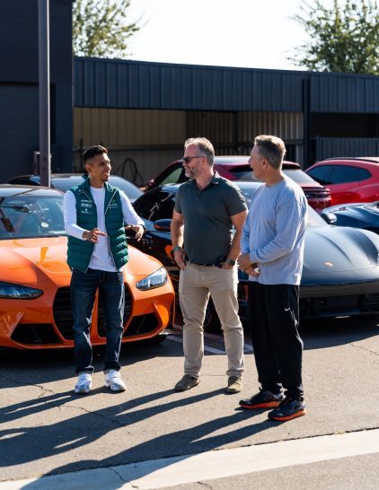 Three men stand outdoors talking near a collection of brightly colored sports cars.