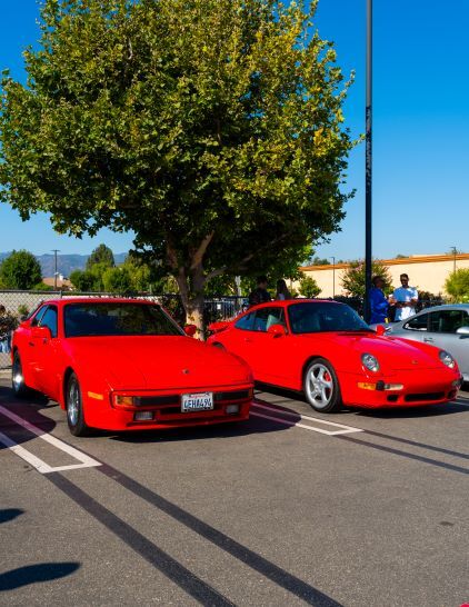 Two red Porsche cars are parked in a lot with a green tree and distant mountains.
