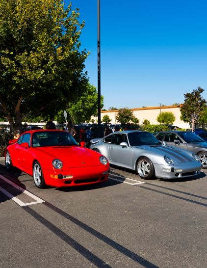 A red and silver Porsche coupe are parked side-by-side in a sunlit parking lot.