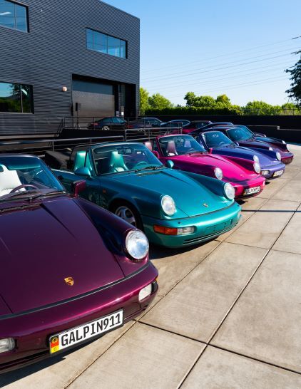 A row of vintage teal, purple, and green Porsche 911 cars are parked outside a modern building.