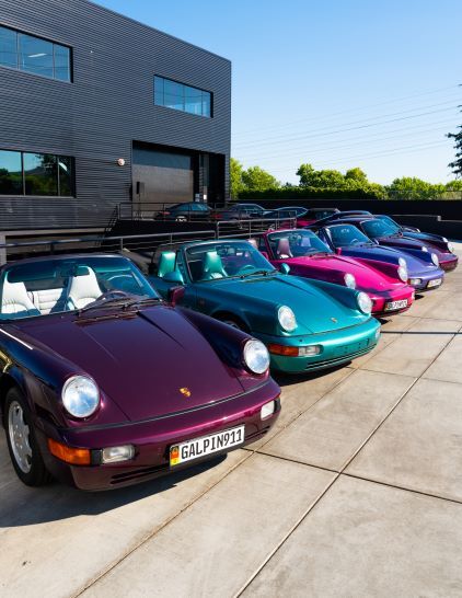 Several vintage Porsche 911 cars in various colors are parked in a row outside a modern building.