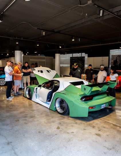 A modified white and green Mazda RX-7 with its hood open draws a crowd of onlookers at an event.