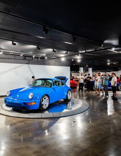A bright blue Porsche 911 sits on a rotating display under spotlights in a modern showroom.