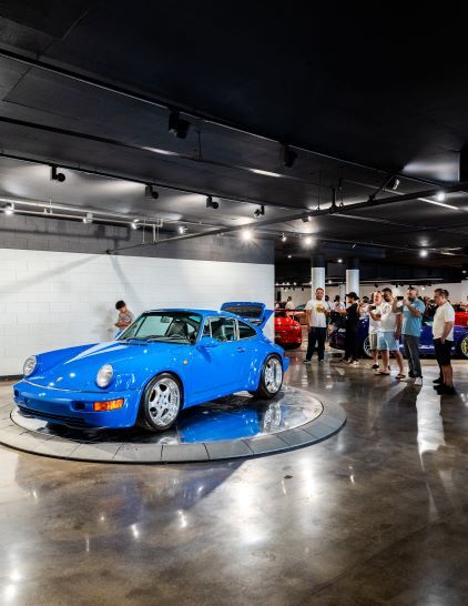 A bright blue Porsche sits on a display turntable as people look at it in an indoor showroom.