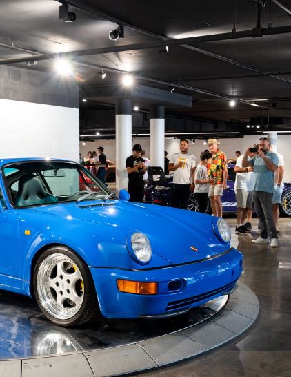 A bright blue Porsche 911 sits on display as people admire it in a modern showroom.
