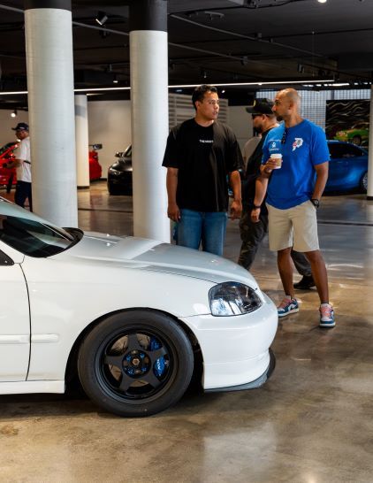Three men converse while standing near a white car in a modern showroom space.