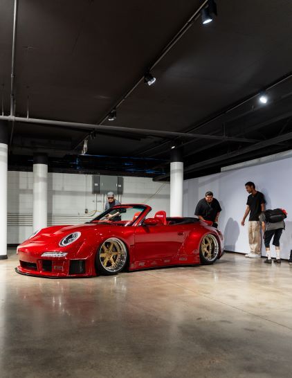 A red Porsche convertible sits on display in a modern showroom with onlookers nearby.
