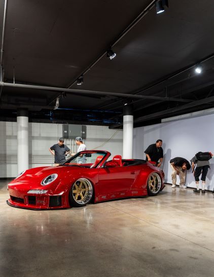 A red Porsche convertible sits on display as several people observe it in a modern showroom.