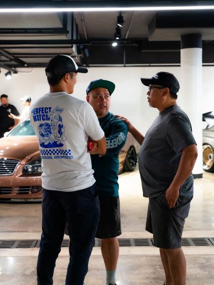 Three men in caps converse in a car showroom with a vehicle visible behind them.