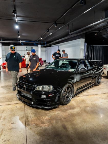 Several people examine a black Nissan Skyline sports car displayed in a large, brightly lit showroom.