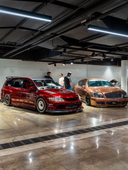 Several customized cars are displayed in a brightly lit, modern parking garage with a small group of people observing them.