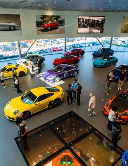 Visitors view a showroom filled with various Porsche cars displayed on a raised platform.