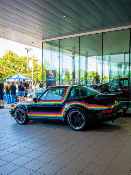 A black Porsche with a rainbow stripe sits inside a glass-walled showroom, surrounded by people.