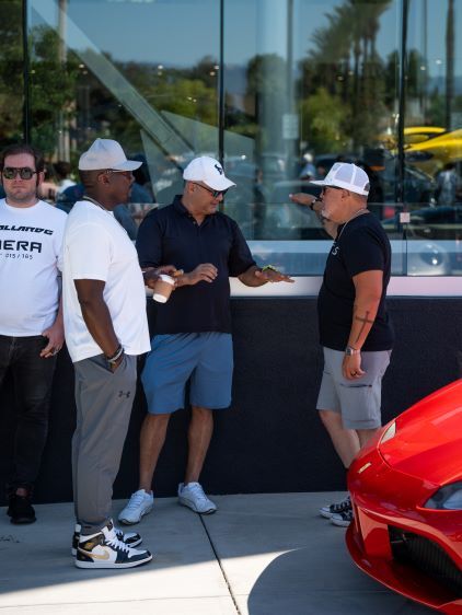 Four men in white caps and casual wear converse in front of a red sports car and glass windows.