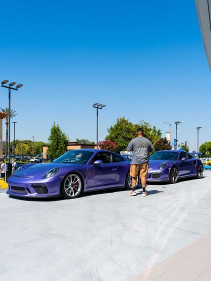 A man walks toward a vibrant purple Porsche parked in a sunny outdoor lot.