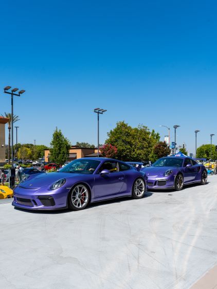 Two purple Porsche cars are parked in a row at an outdoor car show on a sunny day.