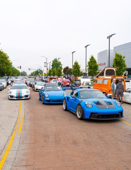 Several blue and silver Porsche sports cars lead a parade down a brick-paved street.