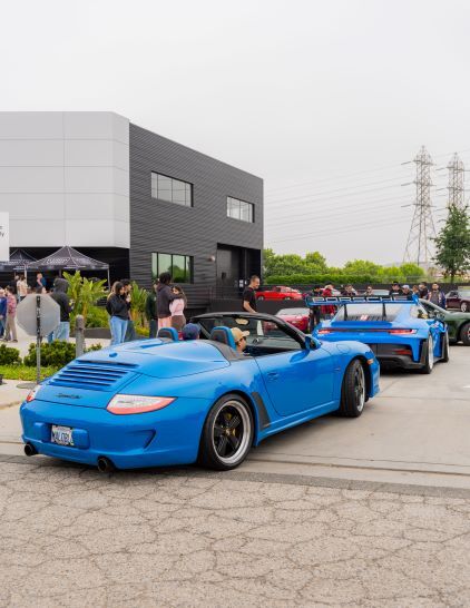 A bright blue Porsche convertible sits parked at a car show with a crowd of people nearby.