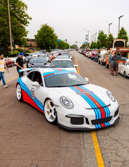 White Porsche 911 GT3 with blue, red, and light blue Martini racing stripes and the number '05' on the side, parked on a brick street with many other cars.