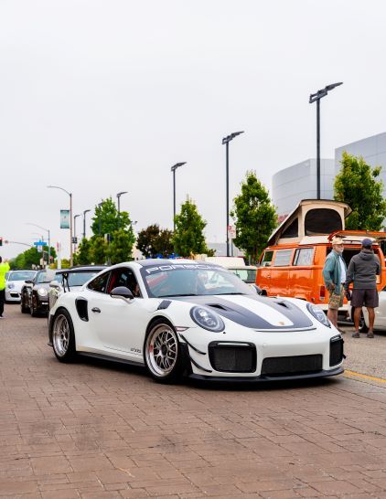 White Porsche GT2RS with a black racing stripe on a brick street, with an orange campervan and people in the background.