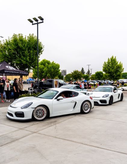 Two white Porsche sports cars parked in an outdoor lot, with trees, tents, and people in the background.