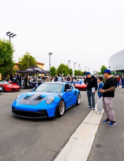 Several people examine a bright blue Porsche sports car at an outdoor car show.