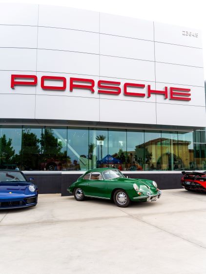 A green vintage Porsche sits in front of a modern Porsche dealership with a blue Porsche nearby.