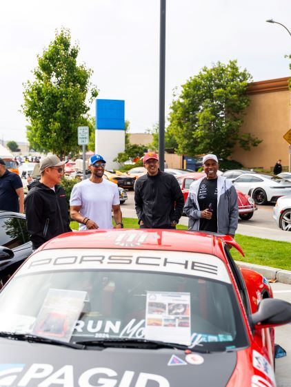 Four men smiling and looking at a red Porsche with 'PORSCHE' written on its windshield at a car show.